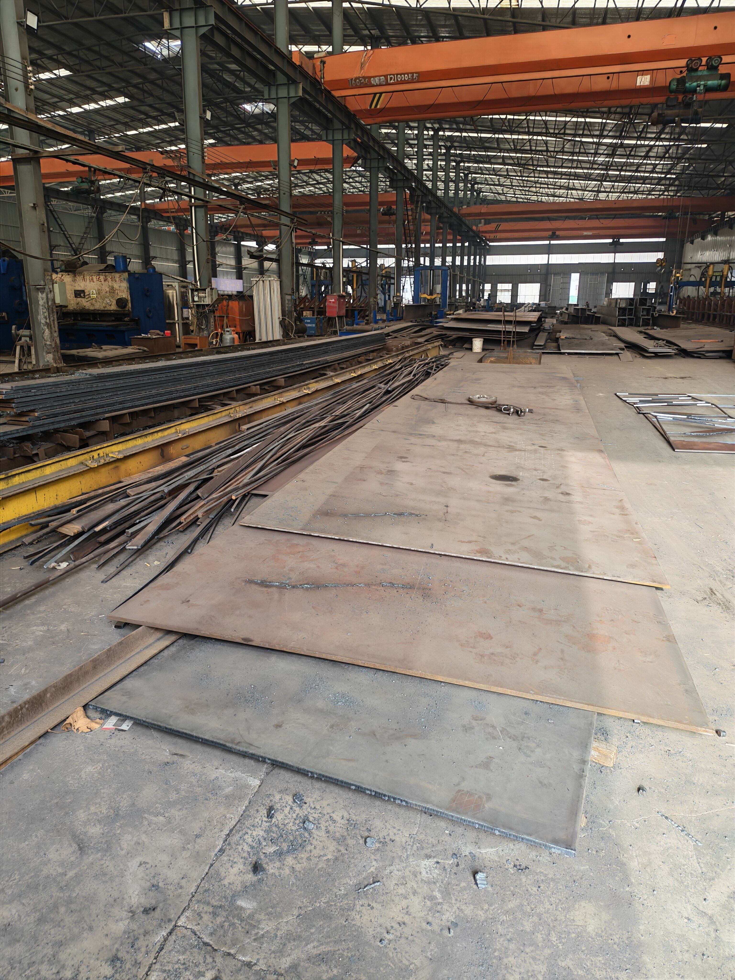 Steel plates stored on the floor of a fabrication workshop beneath an orange overhead crane
