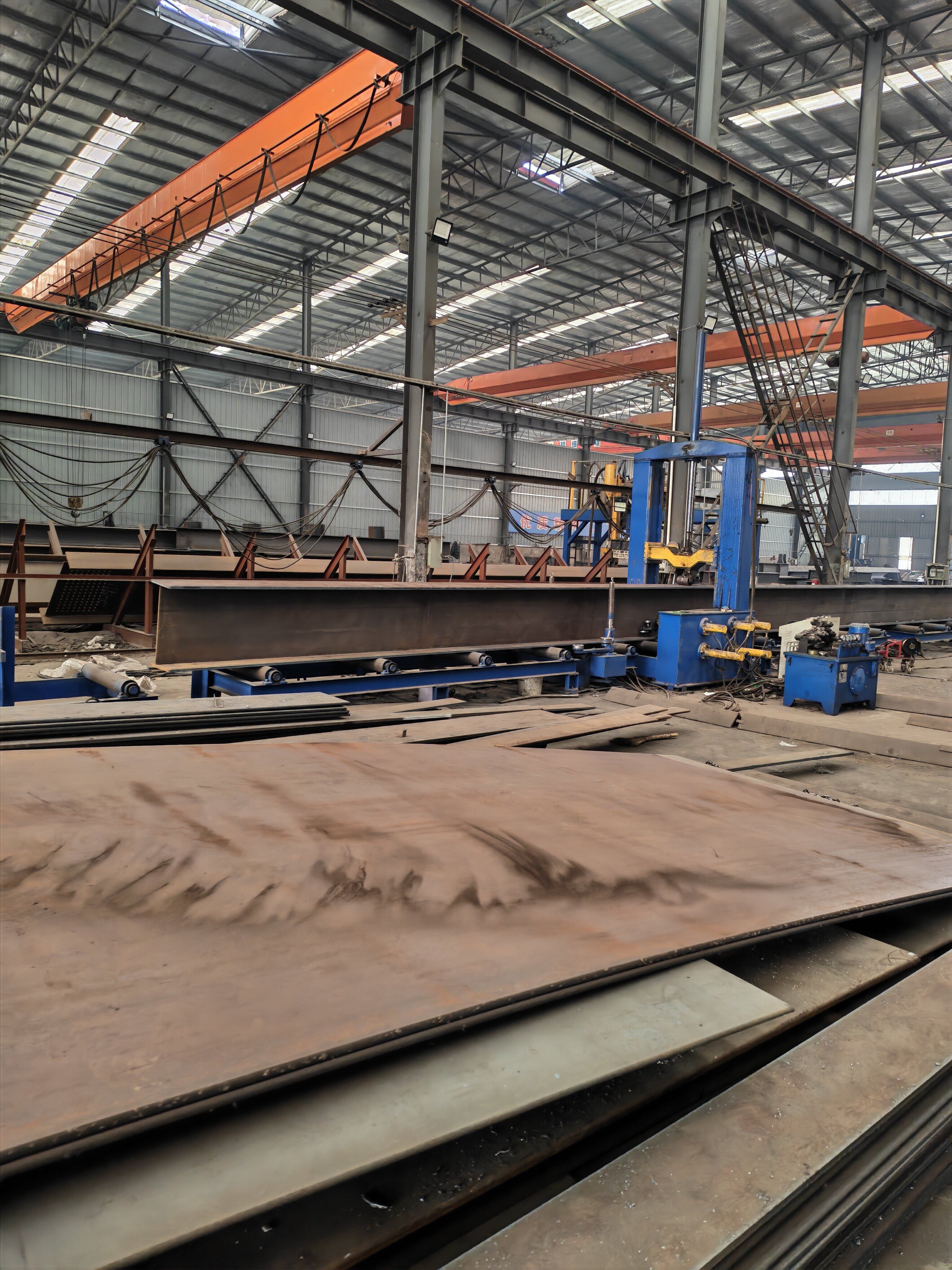 Fabricated steel member on a roller line beside blue processing equipment in a fabrication workshop