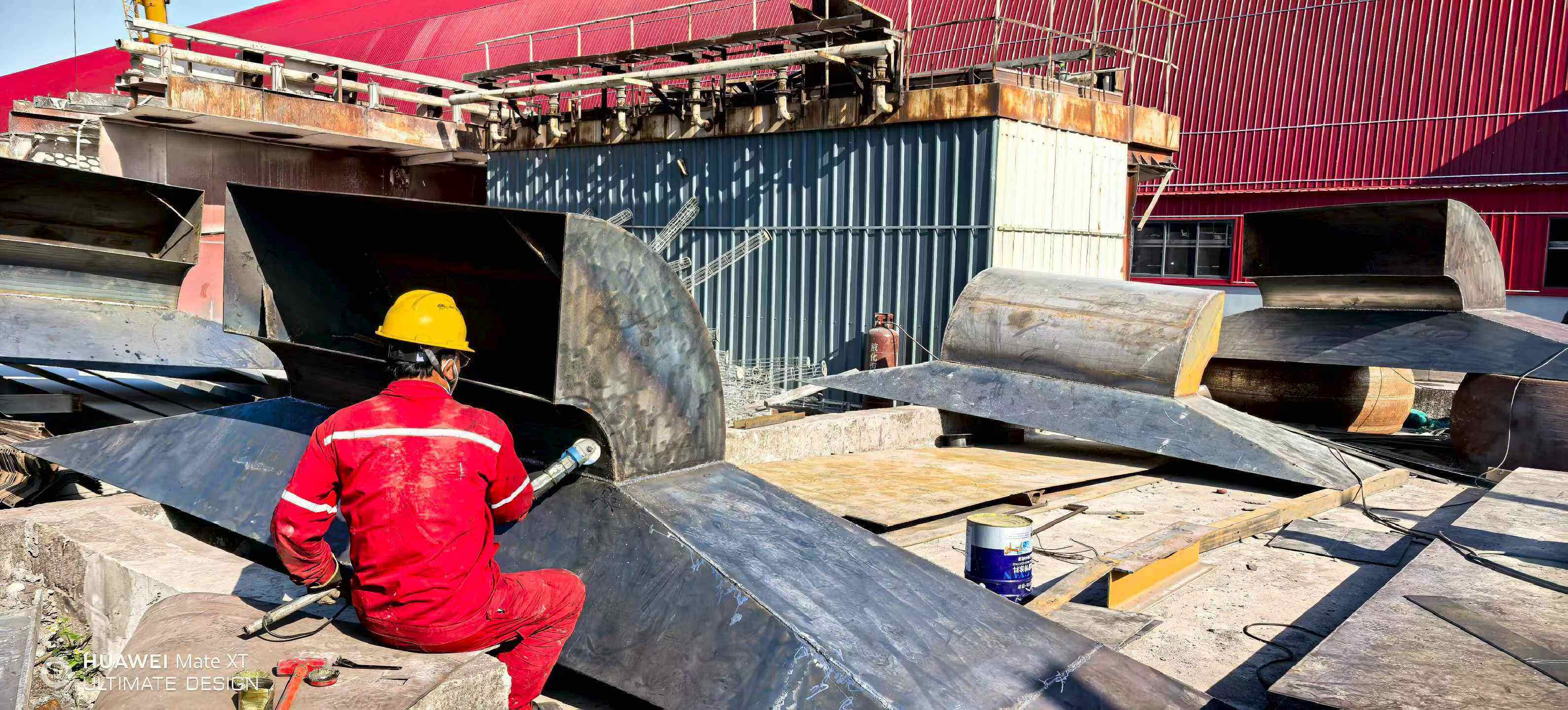 Worker in red uniform grinding a curved steel duct elbow during hood and ductwork fabrication on site