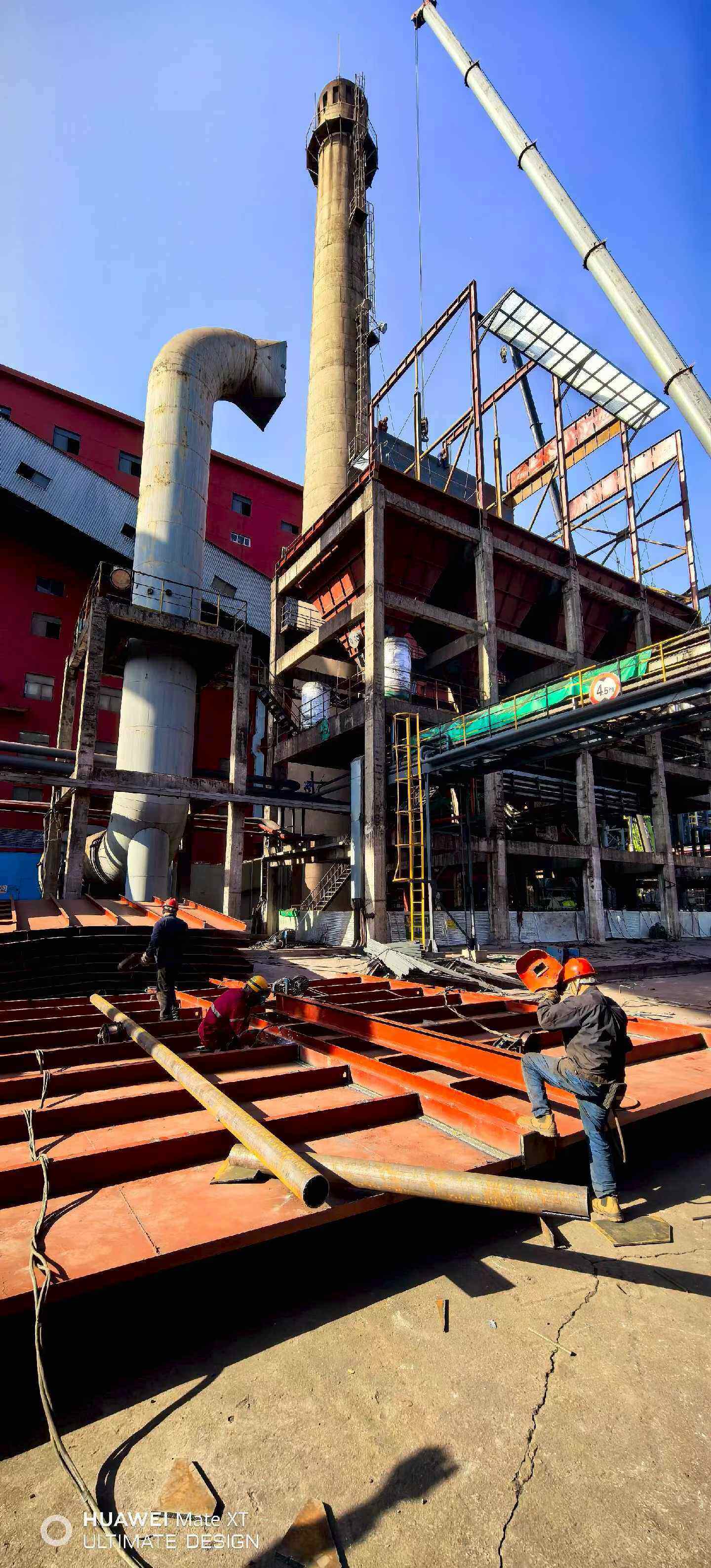 Welders fitting duct floor plates with the main curved flue duct and concrete stack behind at a steel plant