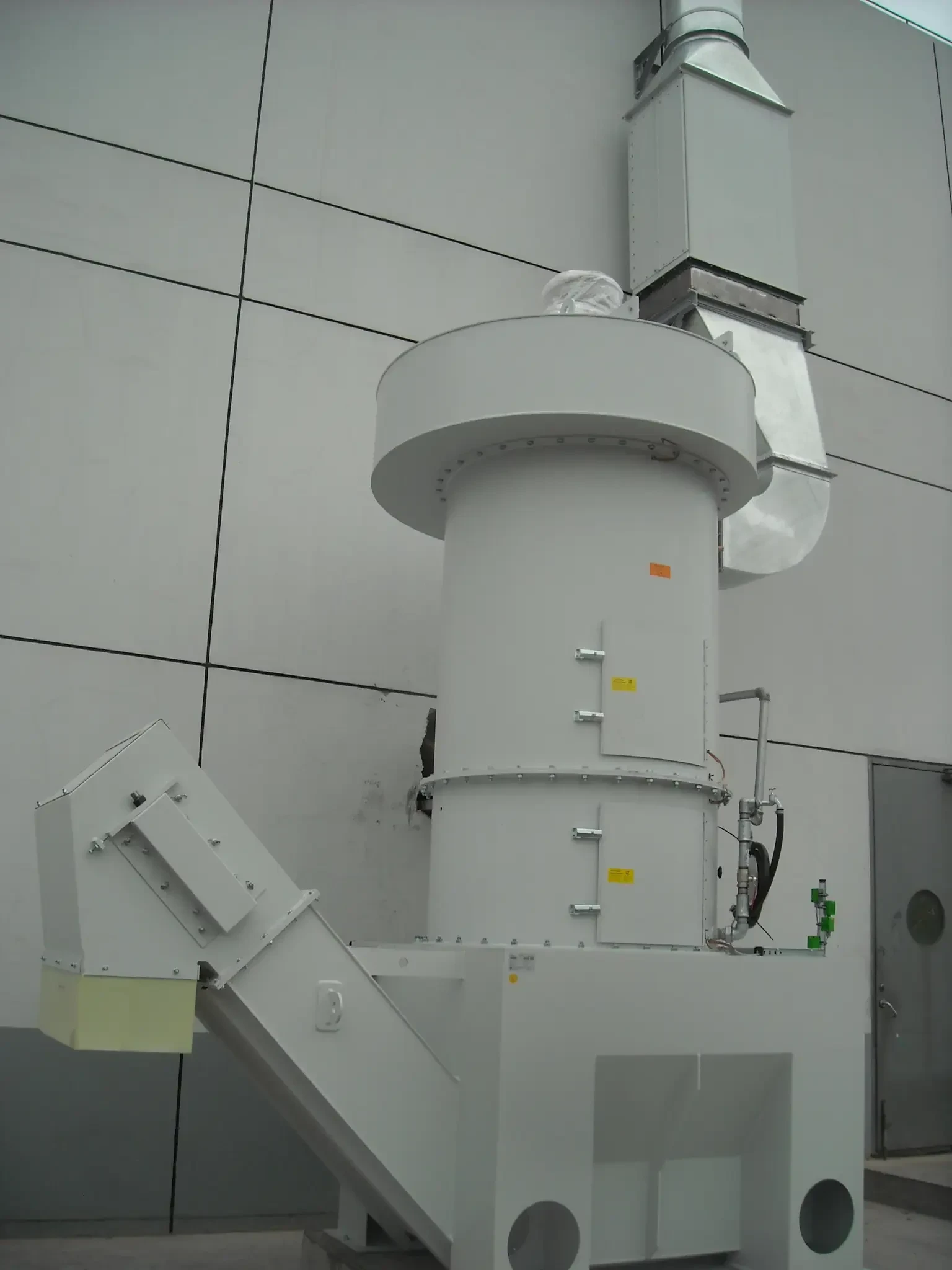 White cylindrical wet dust collector with water supply piping and drain valve installed against a plant wall