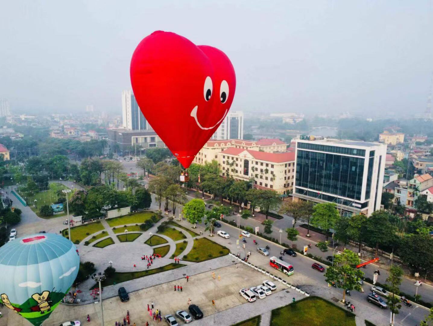 Red Heart-Shaped Hot Air Balloon – Features a smiling face design, flying over the city. Ideal for romantic events and brand promotions.