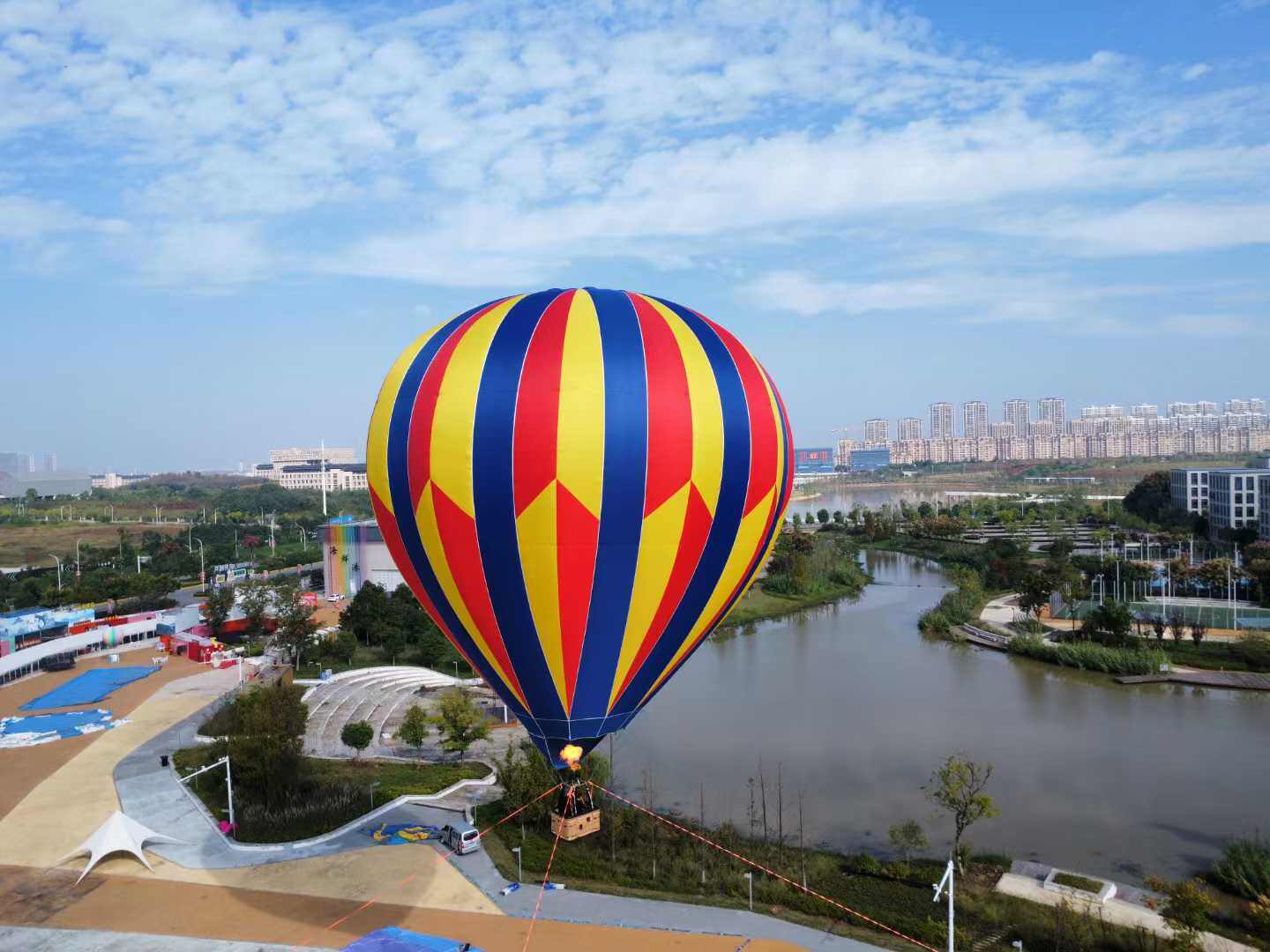 Red, yellow, and blue striped manned hot air balloon aerial photography – Aerial view of a red, yellow, and blue vertical-striped hot air balloon flying over a city river park. Ideal for scenic area tours, urban events, and aerial promotional campaigns with stunning visuals.