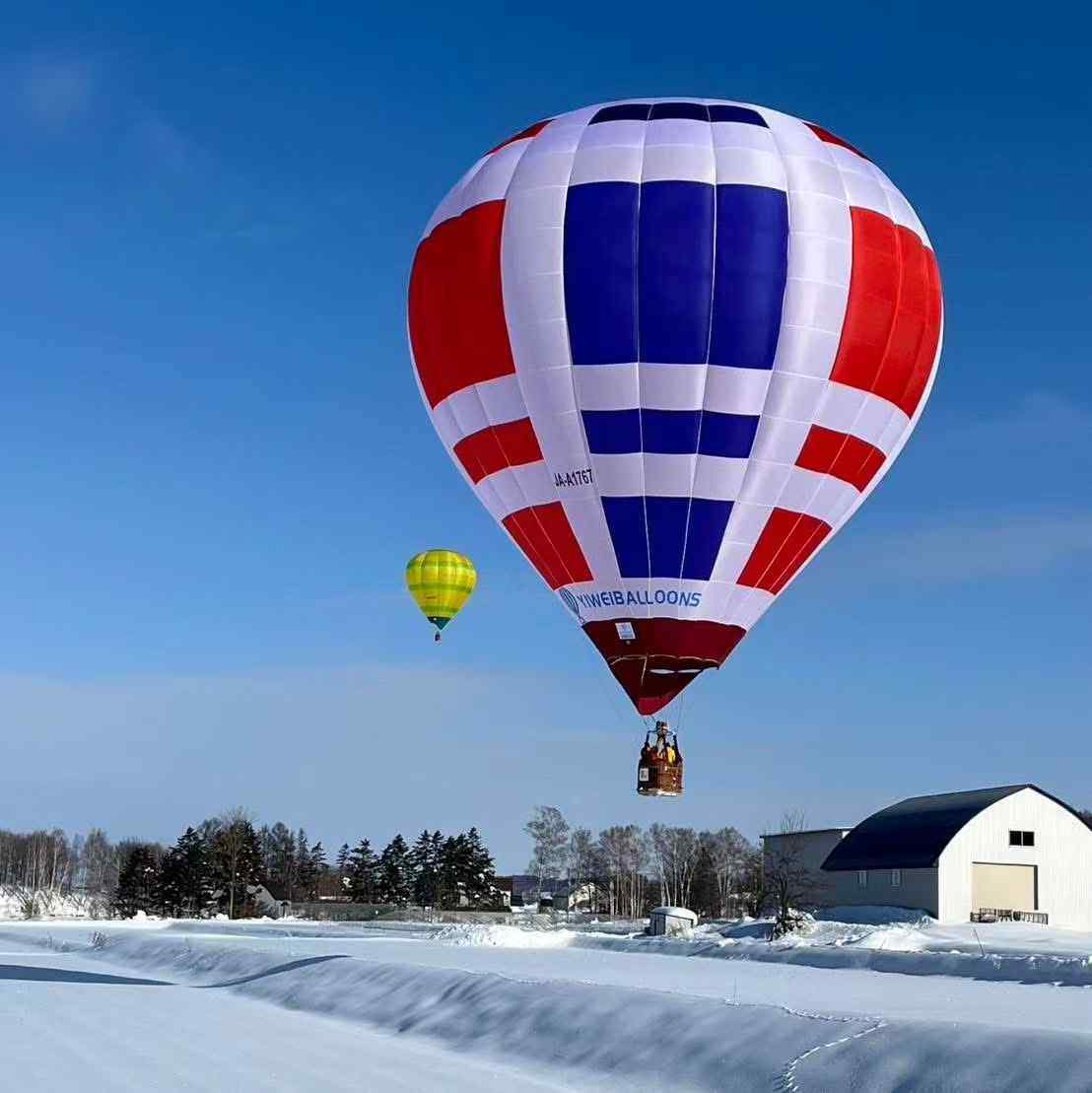 Red White Blue Checkered Manned Hot Air Balloon Flying in Snow - Manned Hot-Air Balloons by Yiwei Balloons - Red white and blue checkered Yiwei Balloons manned hot air balloon flying over snowy landscape with small yellow balloon.
