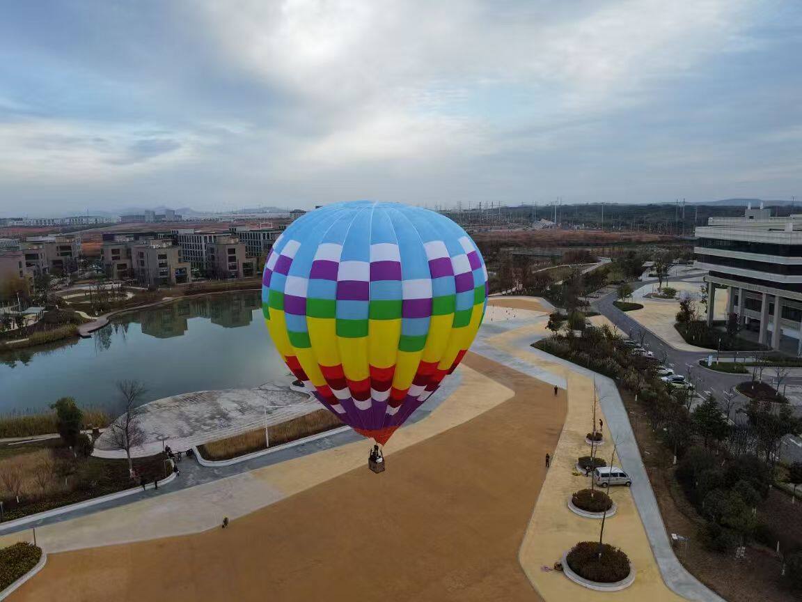 Multi-Color Checkered Manned Hot Air Balloon Aerial View - Manned Hot-Air Balloons by Yiwei Balloons - Aerial drone view of multi-color checkered manned hot air balloon over urban park with lake and buildings.