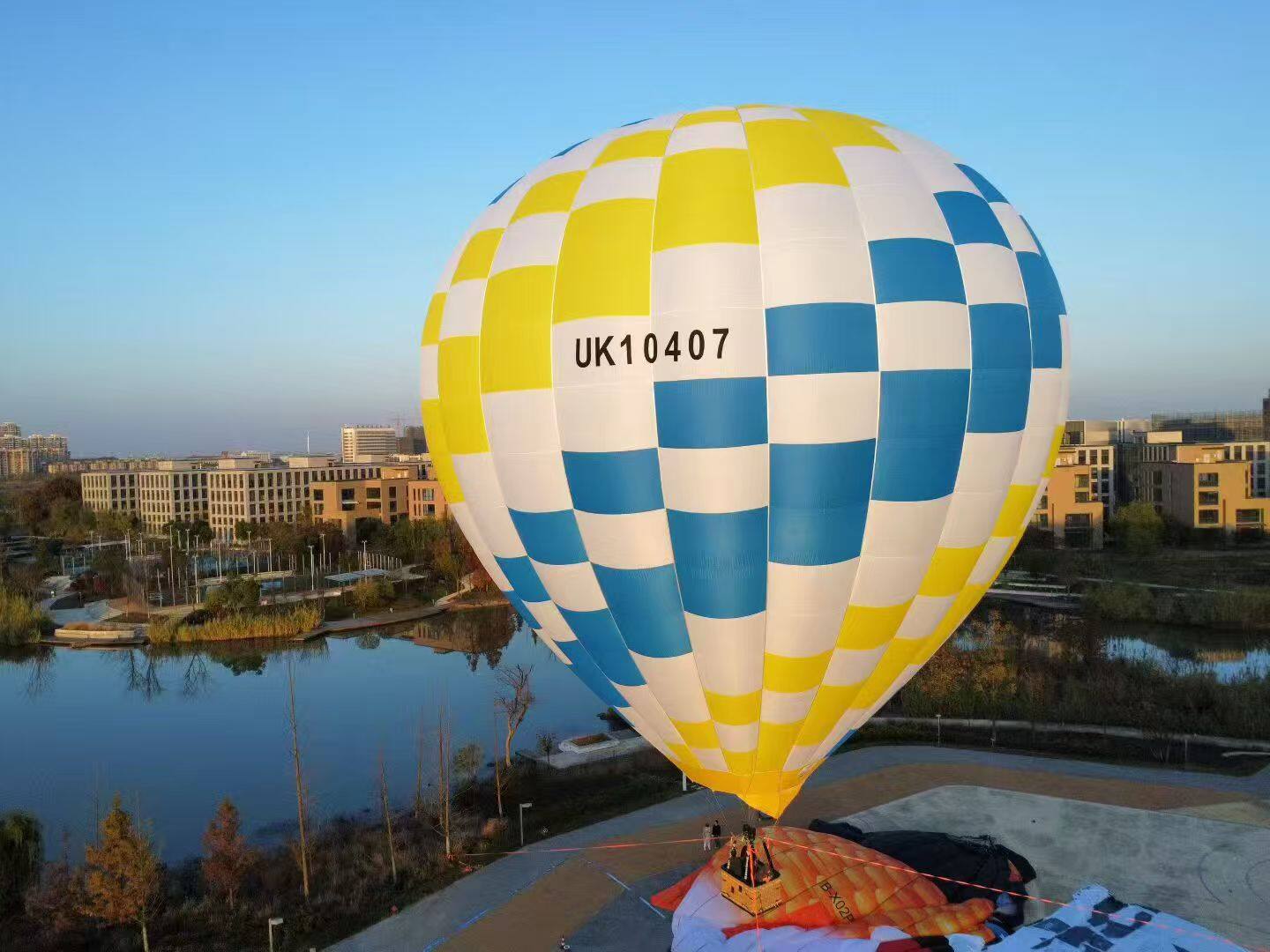 Blue Yellow White Checkered Manned Hot Air Balloon UK10407 - Manned Hot-Air Balloons by Yiwei Balloons - Blue yellow and white checkered manned hot air balloon with registration UK10407 over lake and urban park at sunset.