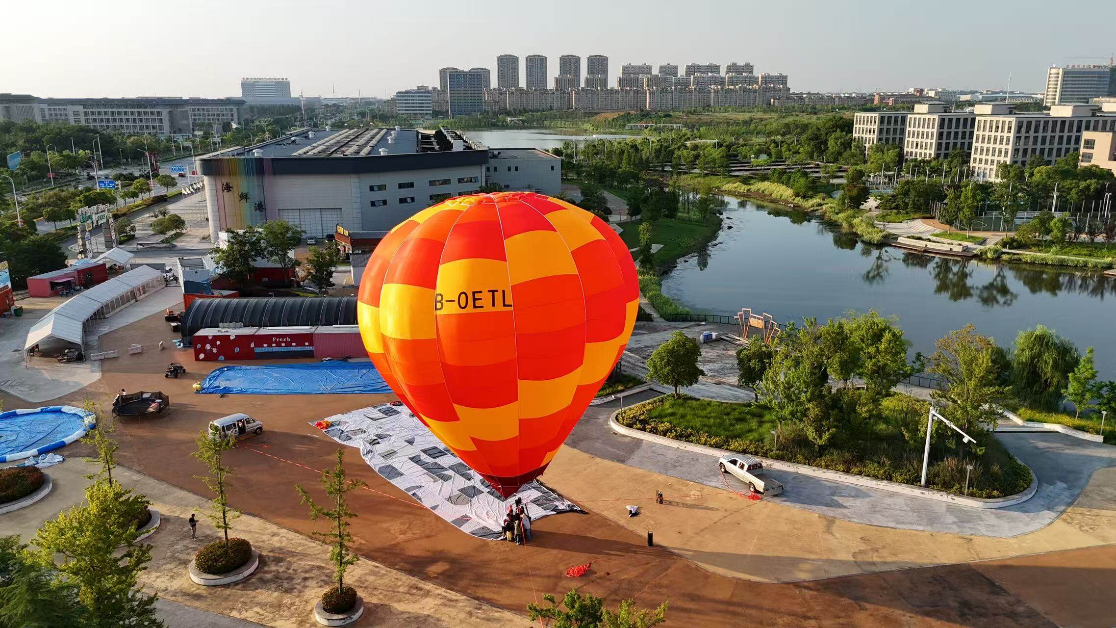 Orange Red Striped Manned Hot Air Balloon Aerial View - Manned Hot-Air Balloons by Yiwei Balloons - Aerial drone view of orange and red striped manned hot air balloon with registration B-OETL over urban park and lake.