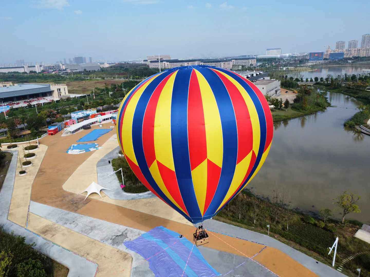 Red Blue Yellow Striped Manned Hot Air Balloon Aerial - Manned Hot-Air Balloons by Yiwei Balloons - Aerial view of red blue and yellow striped manned hot air balloon over lake and urban park with wicker basket.