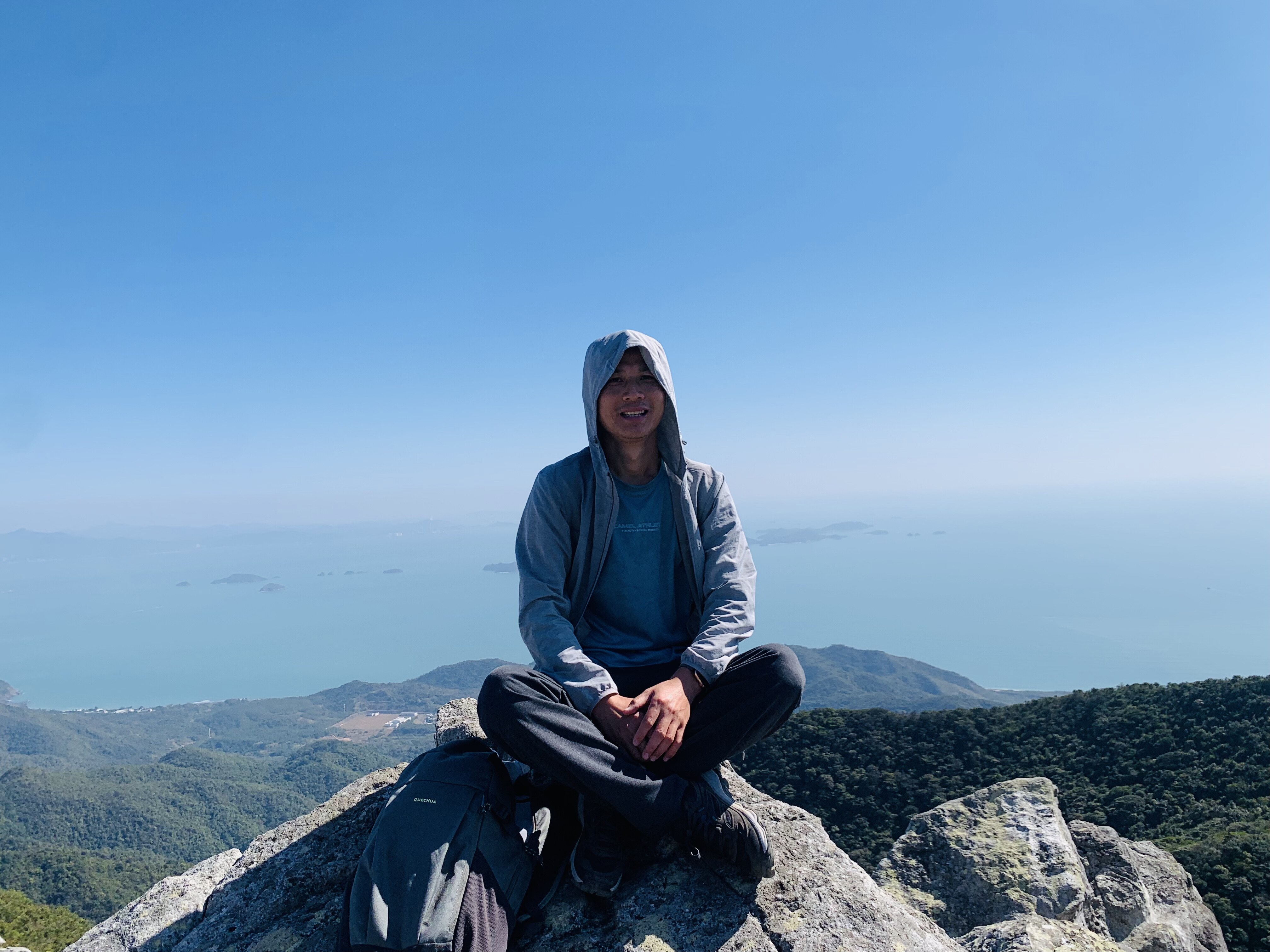 Jacky Liu sitting cross-legged on mountain summit with sea and islands behind