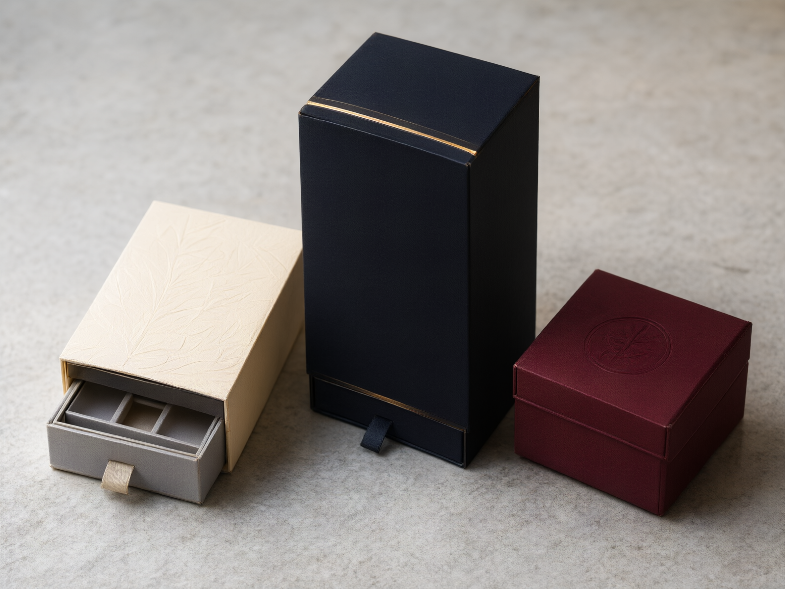 Three luxury drawer boxes of different sizes and finishes arranged on a pale stone surface — matte black perfume box, cream cosmetics box, and deep burgundy gift box