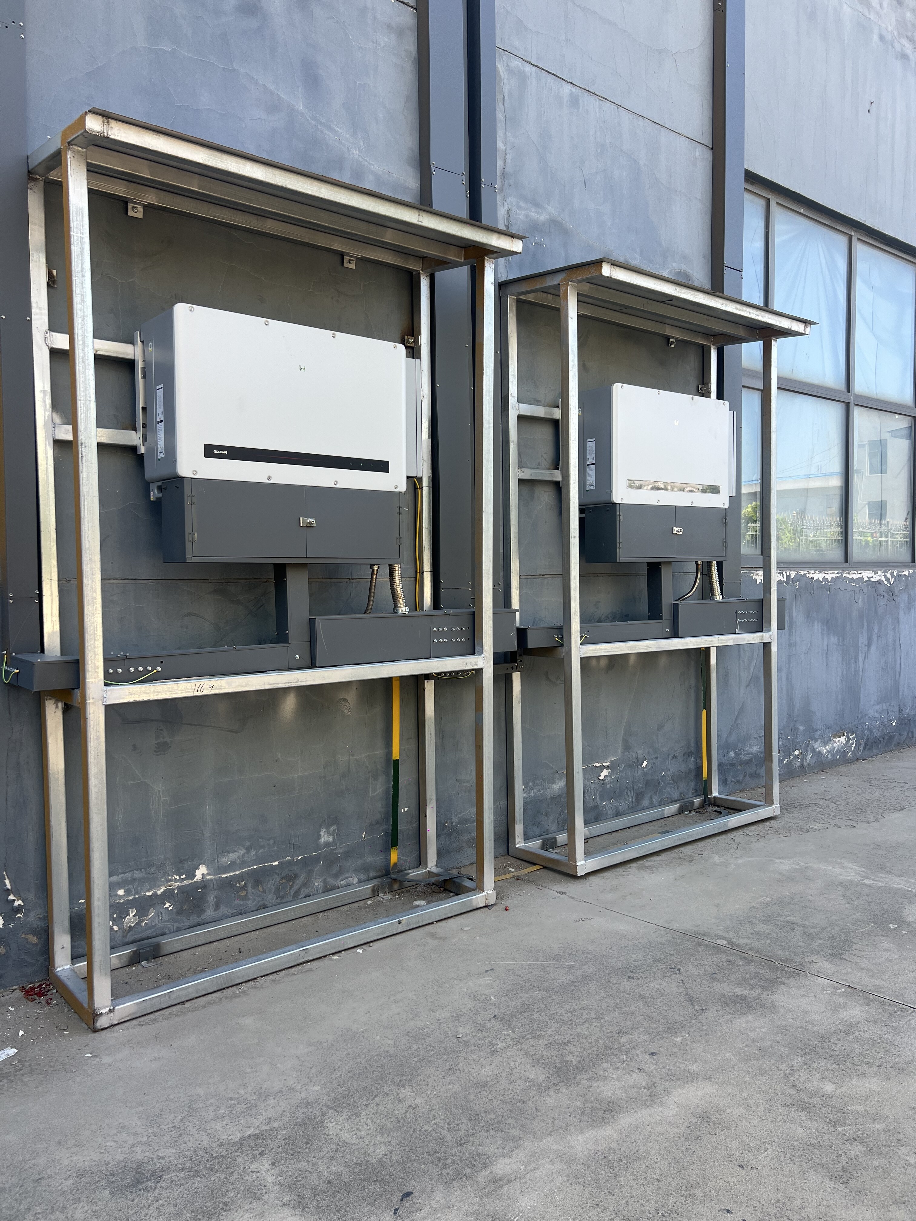 Two white solar inverters mounted on metal frames against gray building exterior wall