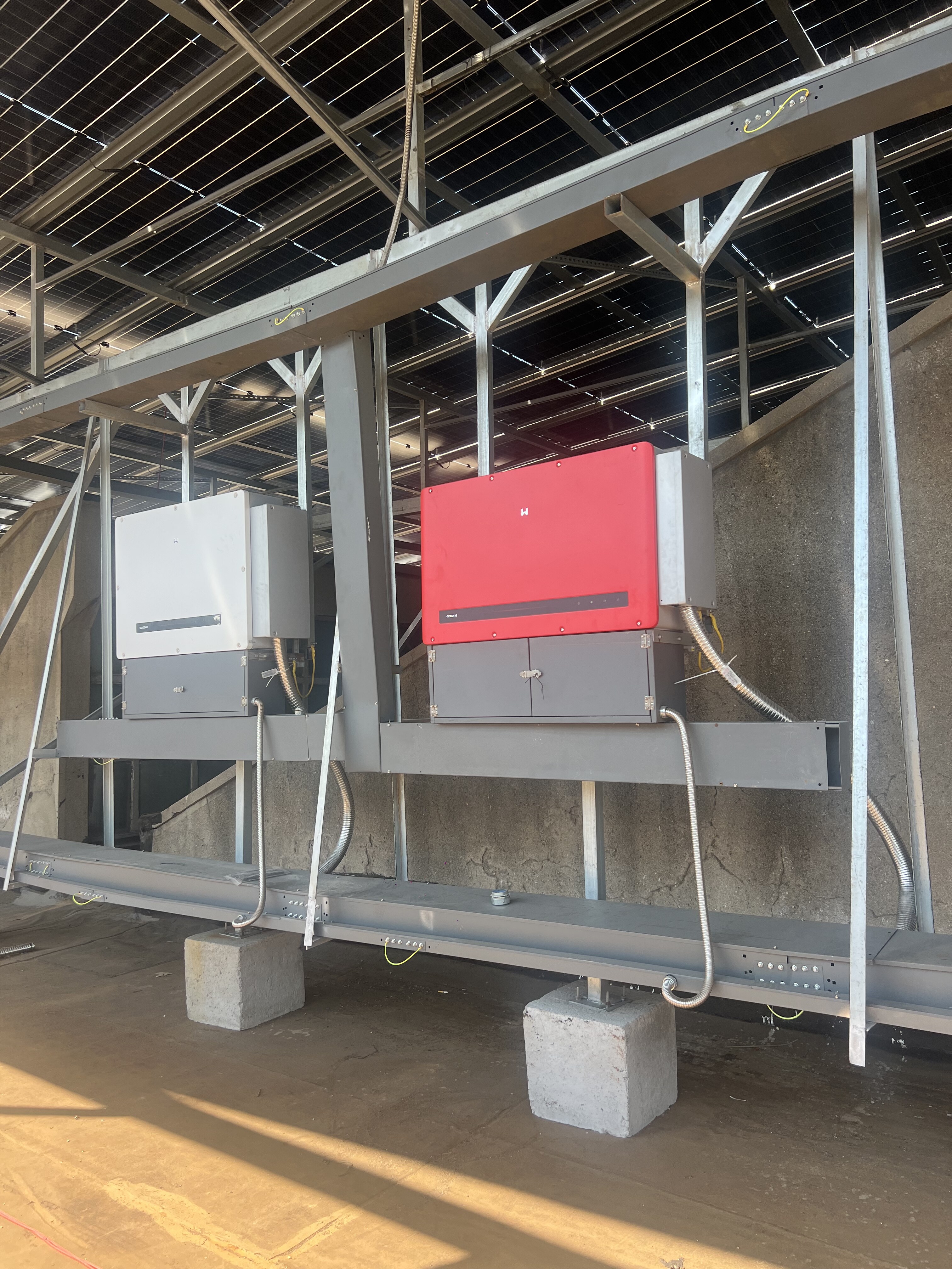 Red and white solar inverters installed under solar panel array with metal support structure