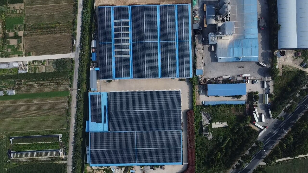 Top-down aerial view of factory buildings with blue roofs and solar panels surrounded by farmland