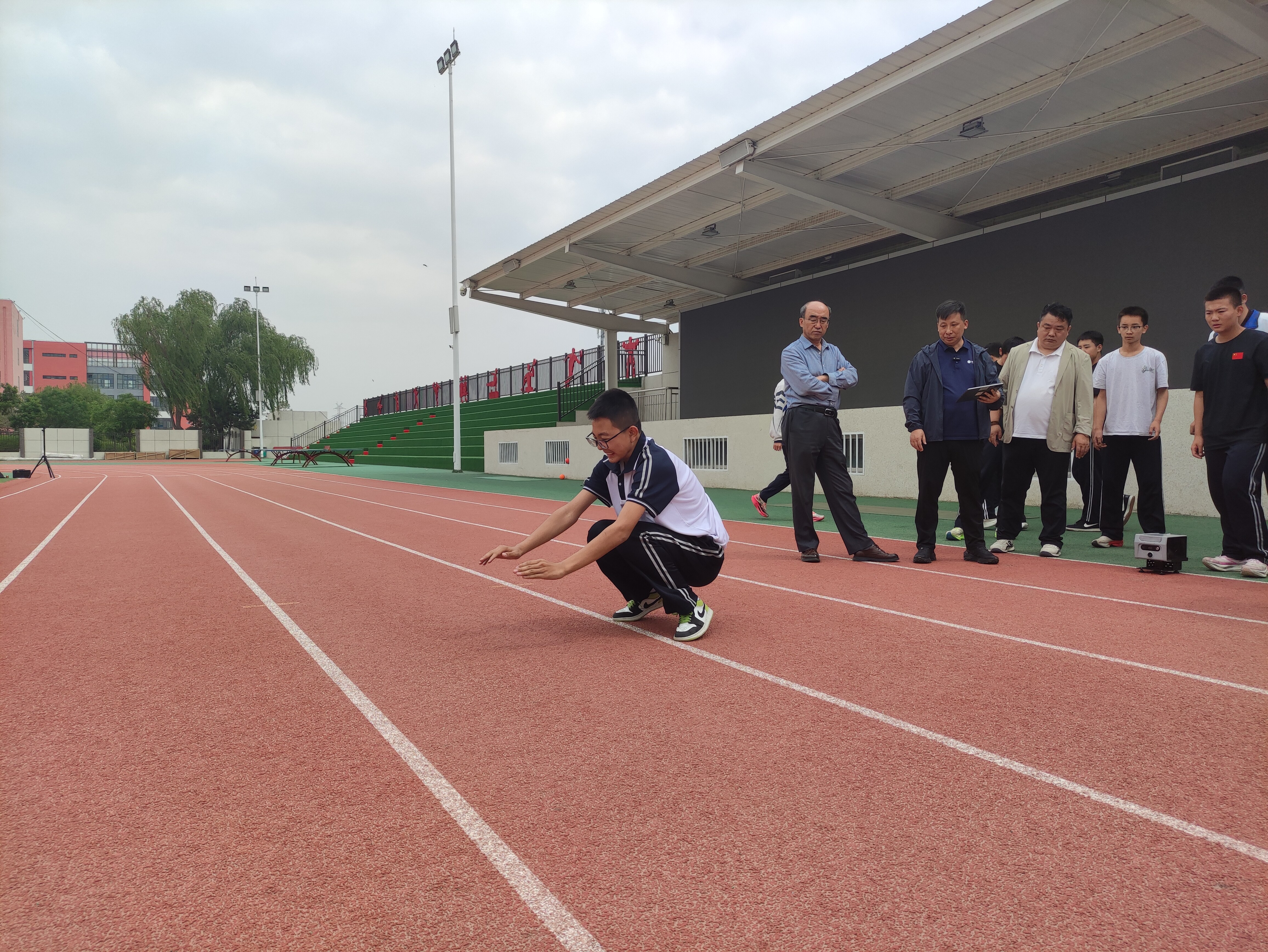 学生在跑道上进行蹲踞式起跑准备，老师和工作人员在旁观察指导