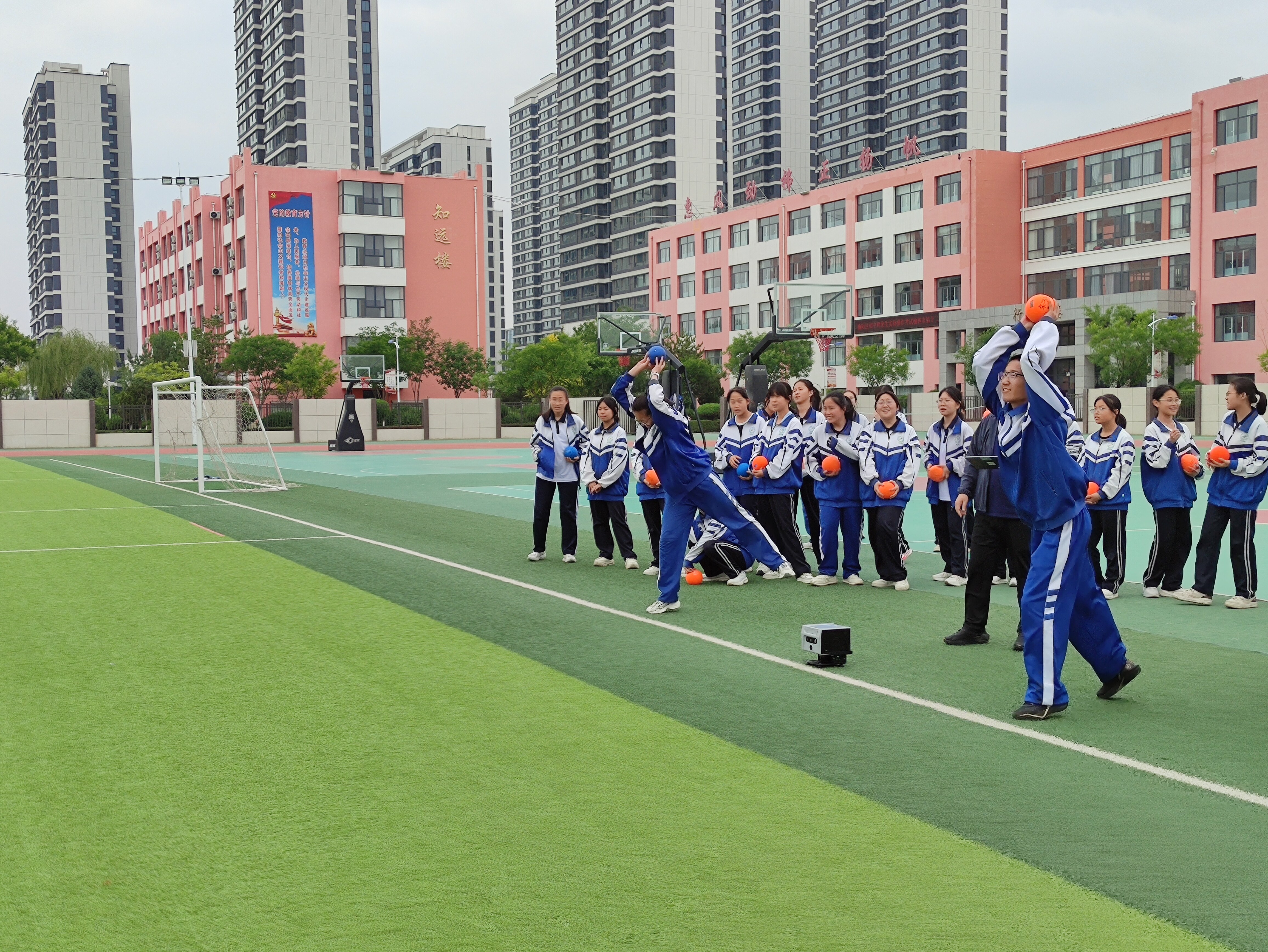 学生在绿色草坪上进行实心球投掷准备动作，同学们排队等候测试